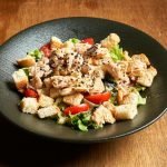 Savory chicken Caesar salad topped with croutons and sesame seeds in a black bowl.