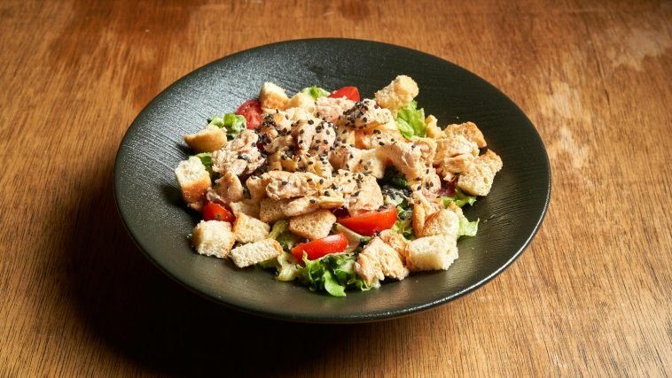 Savory chicken Caesar salad topped with croutons and sesame seeds in a black bowl.
