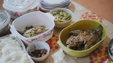 Close-up of a home-cooked meal with fried fish, vegetables, and rice, displayed on a decorated table.