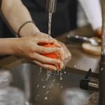 Close-up of hands washing a ripe red tomato under a kitchen faucet. Water splashing in action.