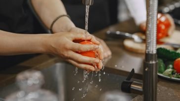 Close-up of hands washing a ripe red tomato under a kitchen faucet. Water splashing in action.