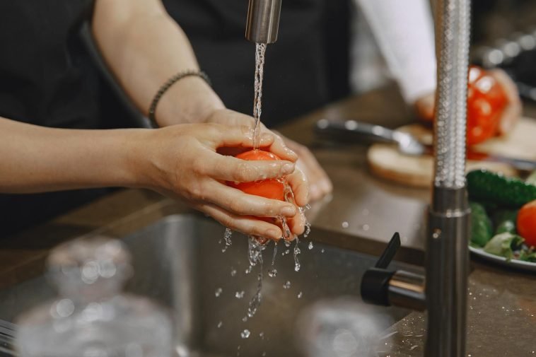 Close-up of hands washing a ripe red tomato under a kitchen faucet. Water splashing in action.