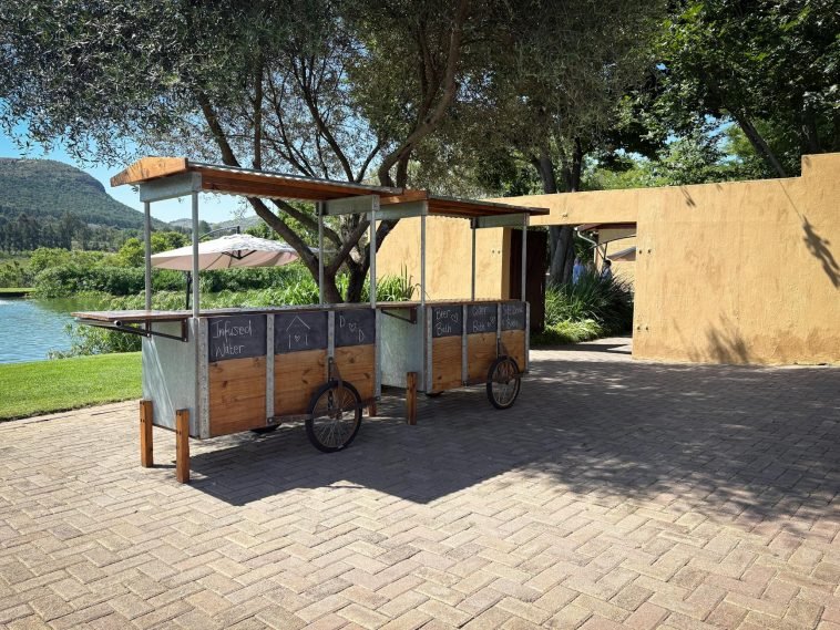 A rustic drink cart set outdoors by a river, offering beverages.