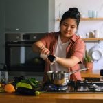 A home cook seasoning a dish on a stove surrounded by fresh vegetables in a modern kitchen.