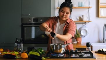 A home cook seasoning a dish on a stove surrounded by fresh vegetables in a modern kitchen.