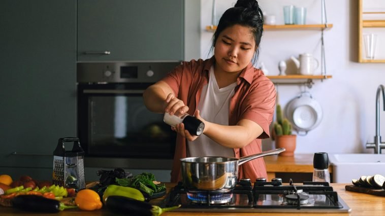 A home cook seasoning a dish on a stove surrounded by fresh vegetables in a modern kitchen.