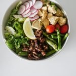 A vibrant salad bowl with asparagus, chicken, radishes, and fresh vegetables on a white surface.
