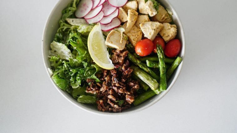 A vibrant salad bowl with asparagus, chicken, radishes, and fresh vegetables on a white surface.