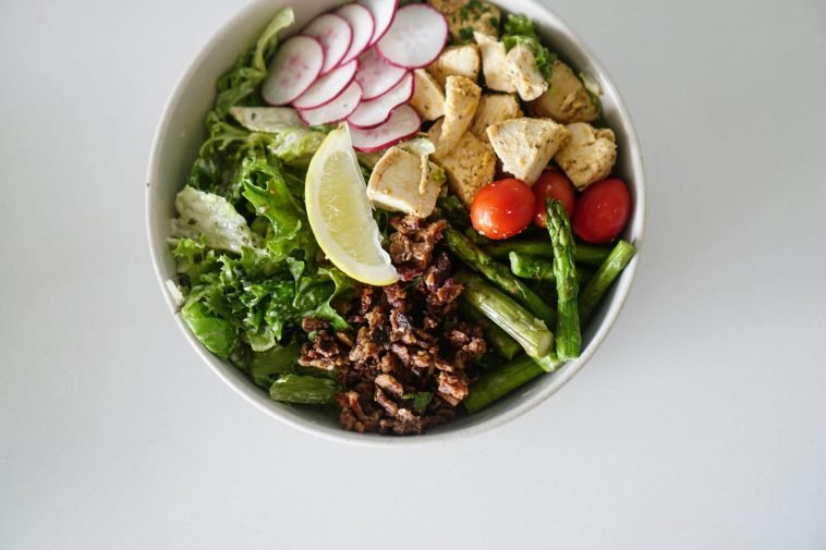 A vibrant salad bowl with asparagus, chicken, radishes, and fresh vegetables on a white surface.