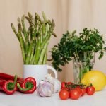 Composition of red cherry tomatoes and peppers with garlics and yellow lemon near white cup with asparagus and green parsley in glass on table