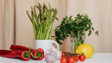 Composition of red cherry tomatoes and peppers with garlics and yellow lemon near white cup with asparagus and green parsley in glass on table
