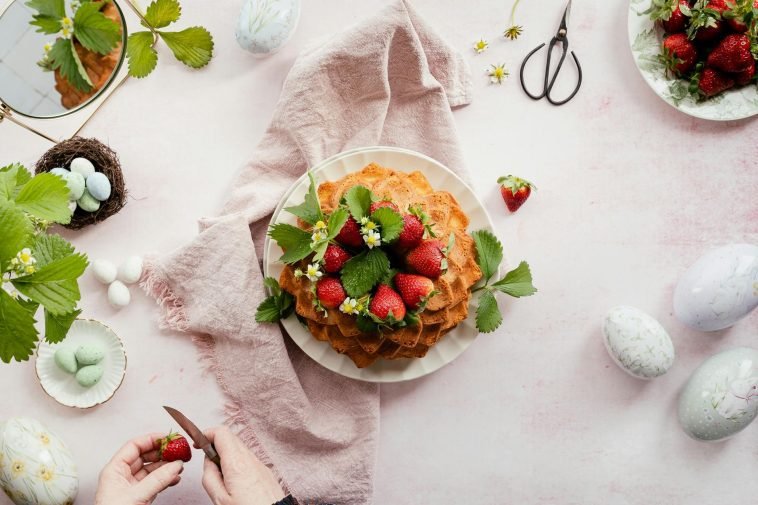 Homemade cake with strawberries on a pink cloth, surrounded by Easter eggs.