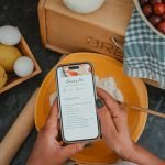 Top view of hands holding a phone with a cherry pie recipe, surrounded by baking ingredients.