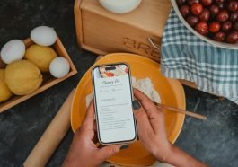 Top view of hands holding a phone with a cherry pie recipe, surrounded by baking ingredients.