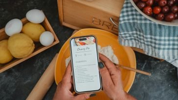 Top view of hands holding a phone with a cherry pie recipe, surrounded by baking ingredients.