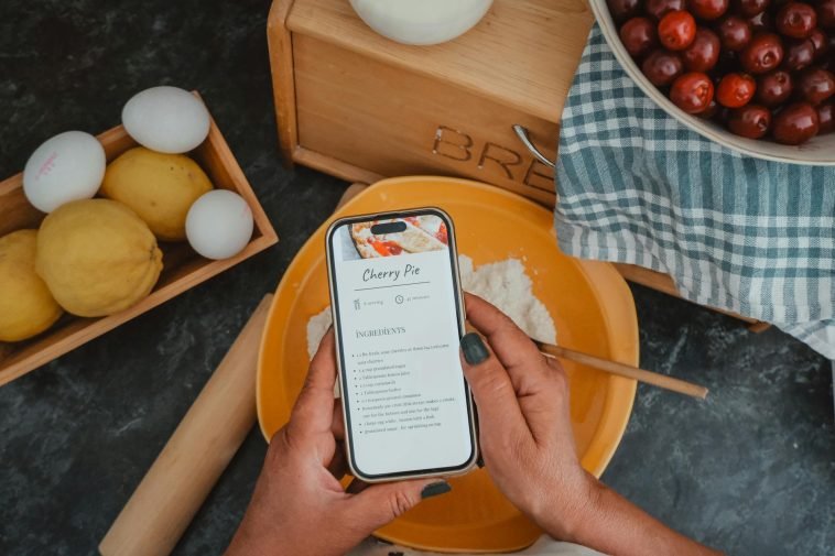 Top view of hands holding a phone with a cherry pie recipe, surrounded by baking ingredients.