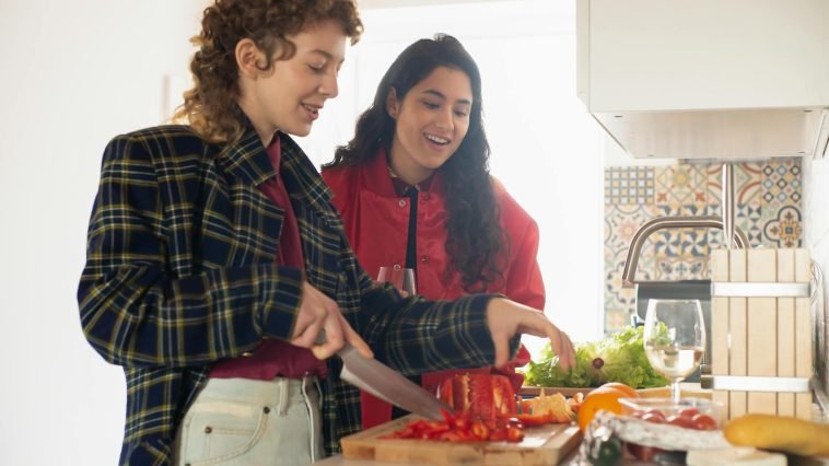Two young women happily preparing a healthy meal with fresh vegetables in a bright kitchen setting.
