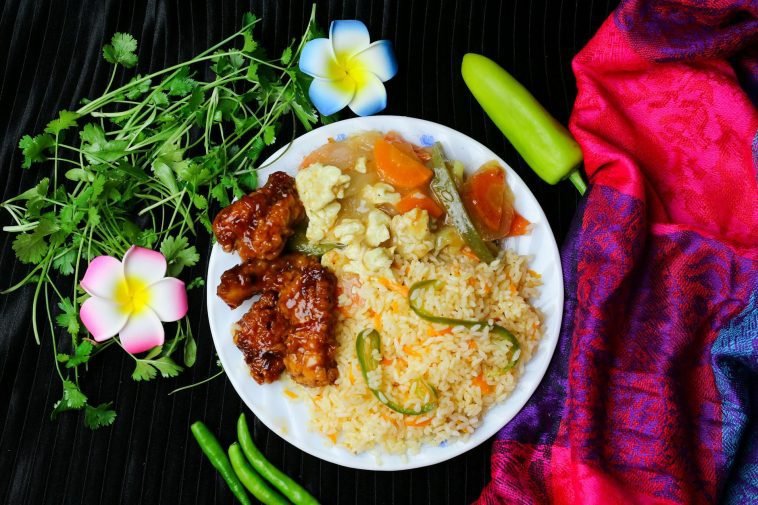 A vibrant dish featuring fried rice with vegetables, glazed chicken, and fresh herbs, garnished with flowers.