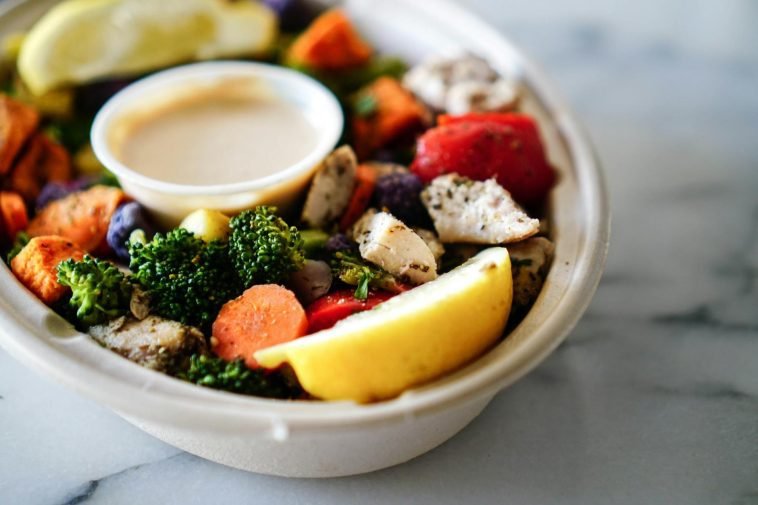 A nutritious bowl of vegetables and chicken served with dipping sauce and lemon slices.
