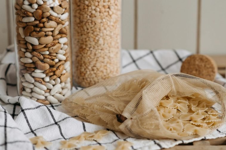Eco-friendly kitchen storage with glass jars of beans and grains beside a mesh bag of pasta on a cloth.