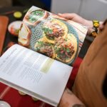 A person holding a cookbook open to a delicious tacos recipe.