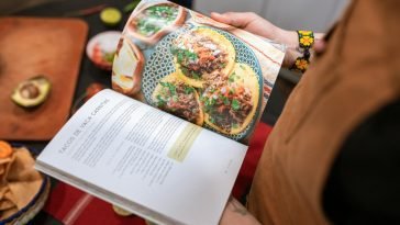 A person holding a cookbook open to a delicious tacos recipe.