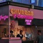 Neon-lit churro and chocolate stand attracting food lovers in an urban market.