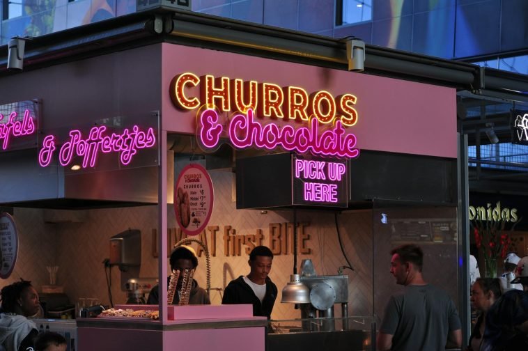 Neon-lit churro and chocolate stand attracting food lovers in an urban market.