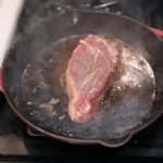Close-up of a steak sizzling in a cast iron skillet on a stove.