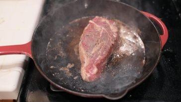 Close-up of a steak sizzling in a cast iron skillet on a stove.