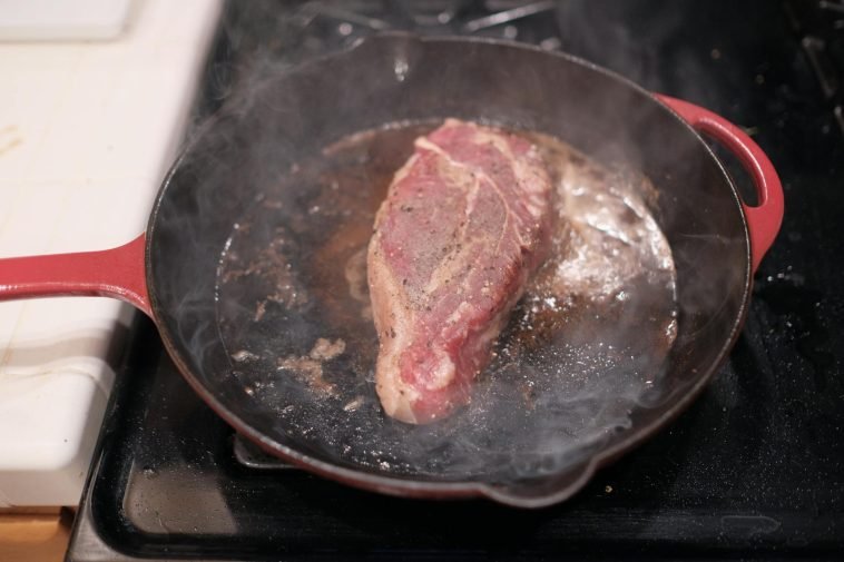 Close-up of a steak sizzling in a cast iron skillet on a stove.