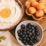 High-angle view of flour with egg, apricots, and blueberries on a wooden table.