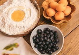 High-angle view of flour with egg, apricots, and blueberries on a wooden table.