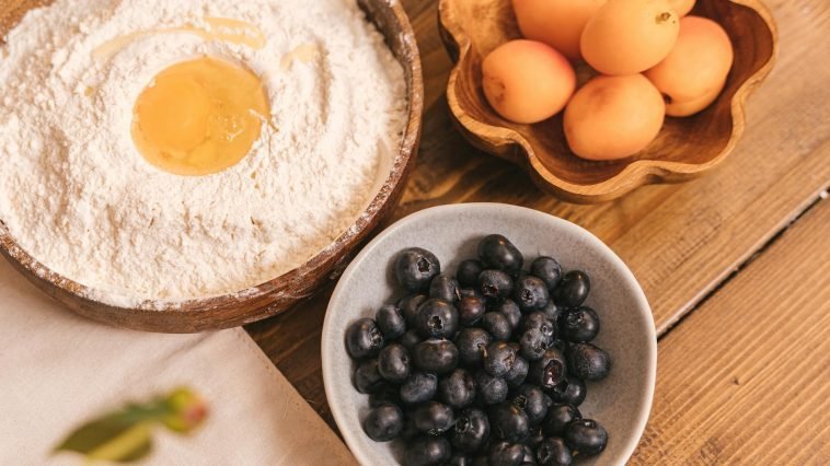 High-angle view of flour with egg, apricots, and blueberries on a wooden table.