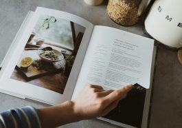 From above of crop faceless woman reviewing article in recipe book in kitchen