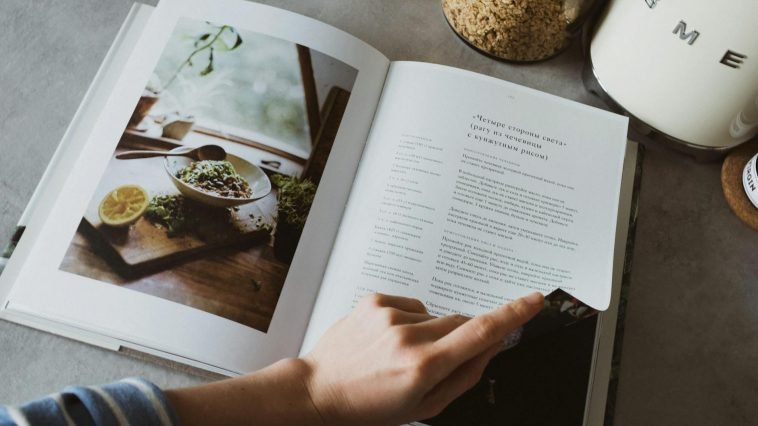 From above of crop faceless woman reviewing article in recipe book in kitchen