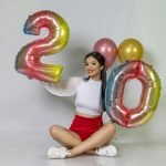 A cheerful woman in a studio celebrating with colorful balloon numbers 20.