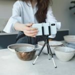 Woman using a smartphone on a tripod for vlogging in the kitchen with bowls.