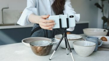 Woman using a smartphone on a tripod for vlogging in the kitchen with bowls.