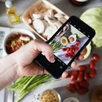 Hand holding smartphone capturing photo of fresh vegetables on a kitchen counter.