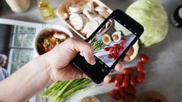 Hand holding smartphone capturing photo of fresh vegetables on a kitchen counter.