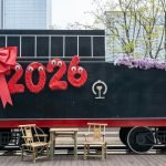 A festive train decorated with '2026' and a red bow, set in an urban environment.