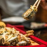 A delicious sushi roll held with chopsticks above a wooden tray in a restaurant setting.