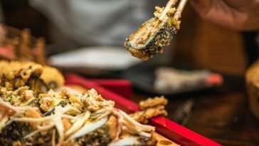 A delicious sushi roll held with chopsticks above a wooden tray in a restaurant setting.