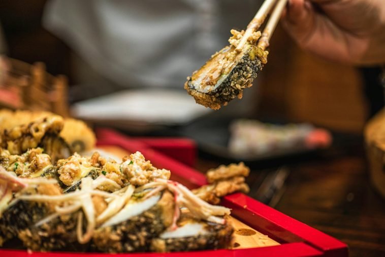 A delicious sushi roll held with chopsticks above a wooden tray in a restaurant setting.