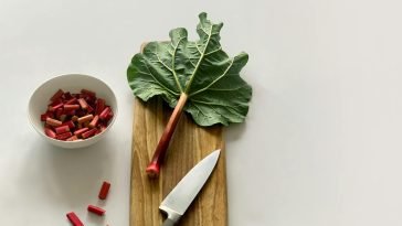Fresh rhubarb stalk and leaves on a wooden cutting board with a knife, ready for cooking.