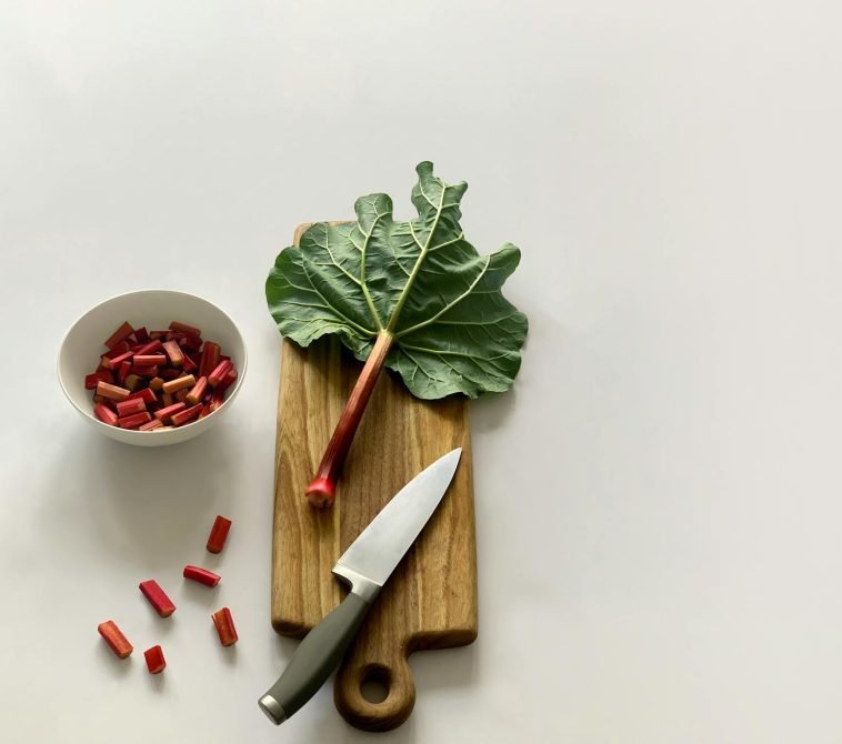 Fresh rhubarb stalk and leaves on a wooden cutting board with a knife, ready for cooking.